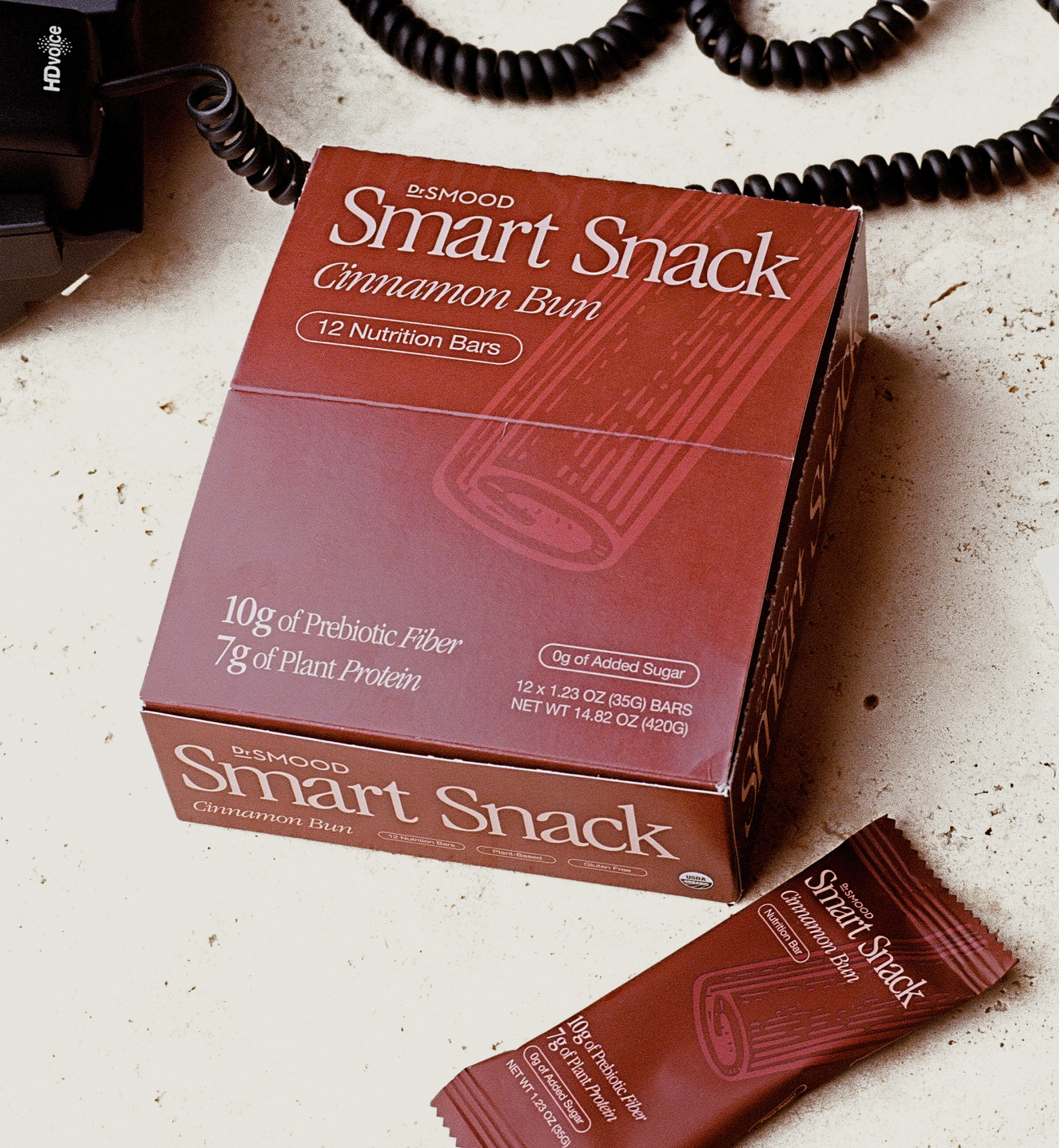 This image showcases the Dr. Smood Smart Snack Cinnamon Bun box of nutrition bars placed on a light-colored surface. The box is dark red and prominently features the product name and key nutritional benefits, including “10g of Prebiotic Fiber” and “7g of Plant Protein,” along with “0g of Added Sugar.” One of the individual bars is also visible in front of the box, highlighting its minimalist design and the cinnamon stick illustration. A telephone cord is seen in the background.