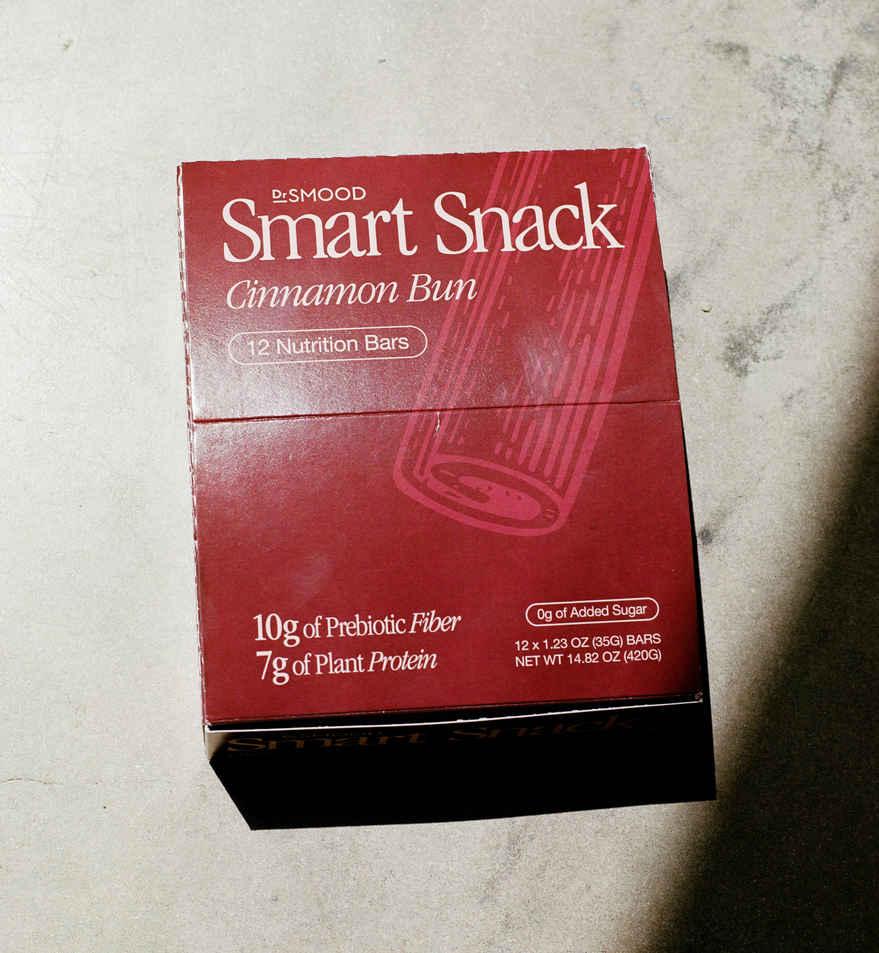 This image shows the Dr. Smood Smart Snack Cinnamon Bun box of nutrition bars placed on a light-colored surface. The box is dark red and prominently features the product name and key nutritional benefits, including “10g of Prebiotic Fiber” and “7g of Plant Protein,” along with “0g of Added Sugar.” The box design also includes an illustration of a cinnamon stick. The light and shadow in the image add a textured look to the surface, creating an interesting visual effect.